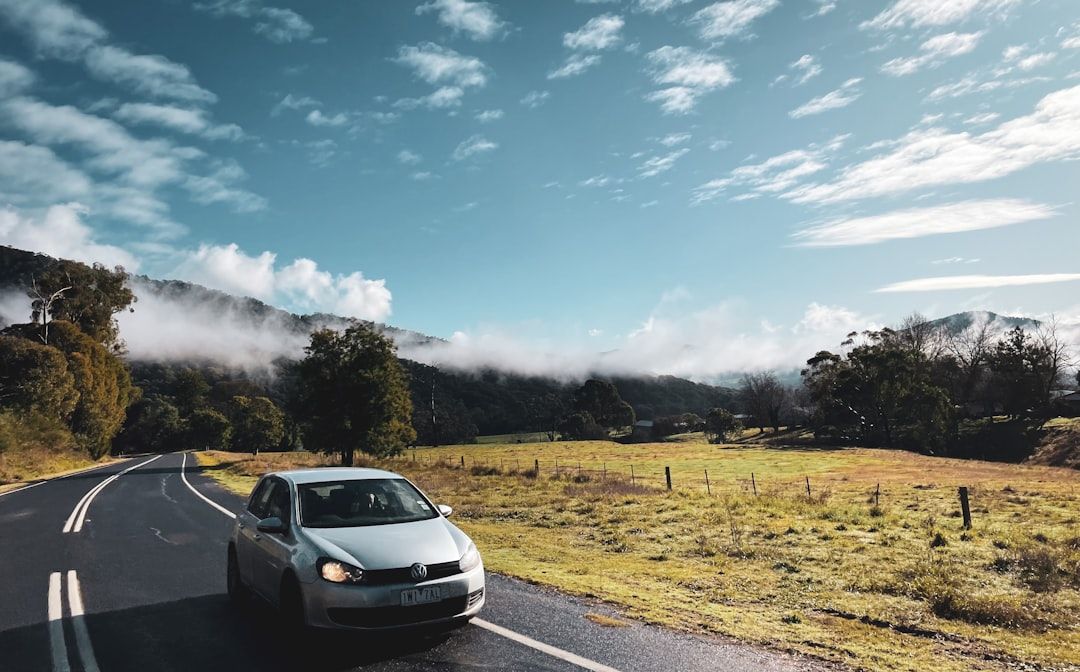 A modern white SUV rental car parked at a scenic lookout point overlooking a vast, sun-drenched Australian landscape with a c